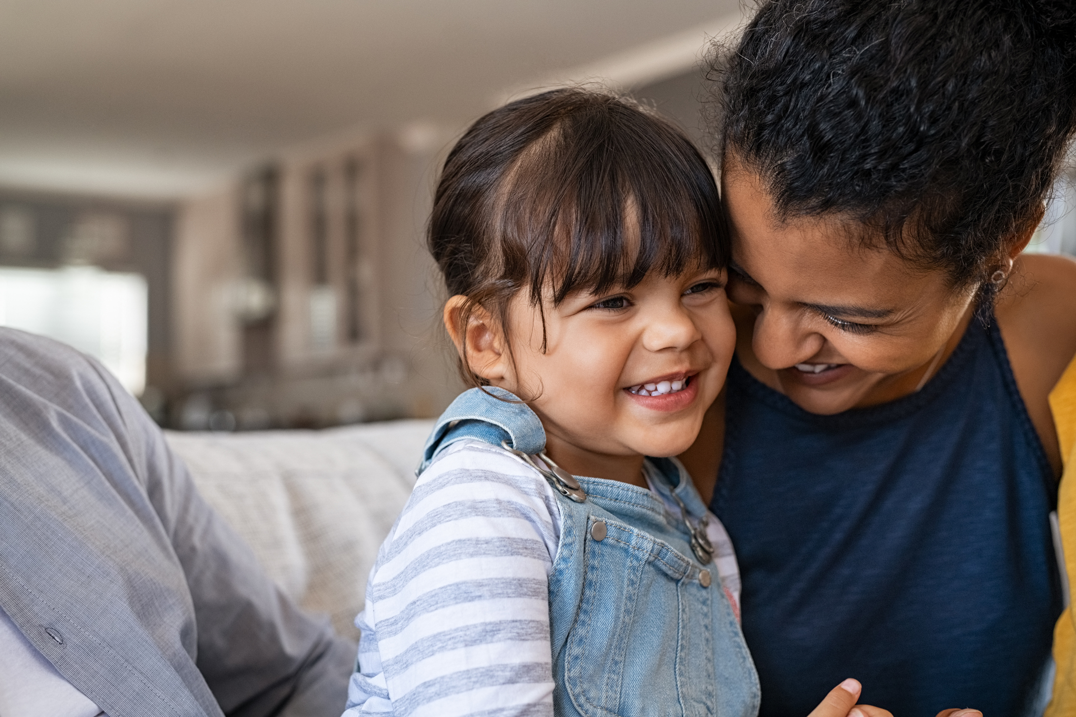 Beautiful black mother embracing little girl sitting on couch with copy space. Cute daughter hugging african american mother and smiling together. Happy latin mom jokes with her kid at home and playing together.